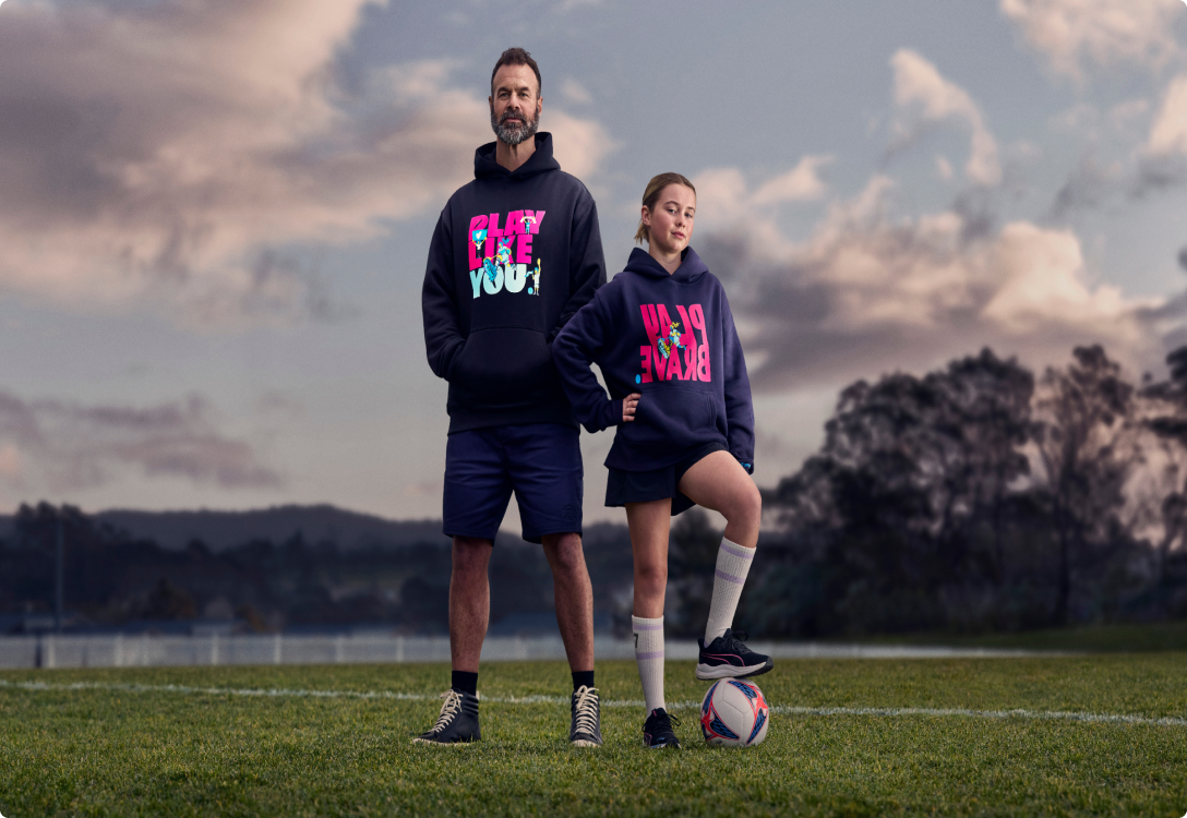 SupportHER Merch - Support Image of a girl playing football wearing the Play Brave hoodie and her father supporting her wearing the Play Like You hoodie